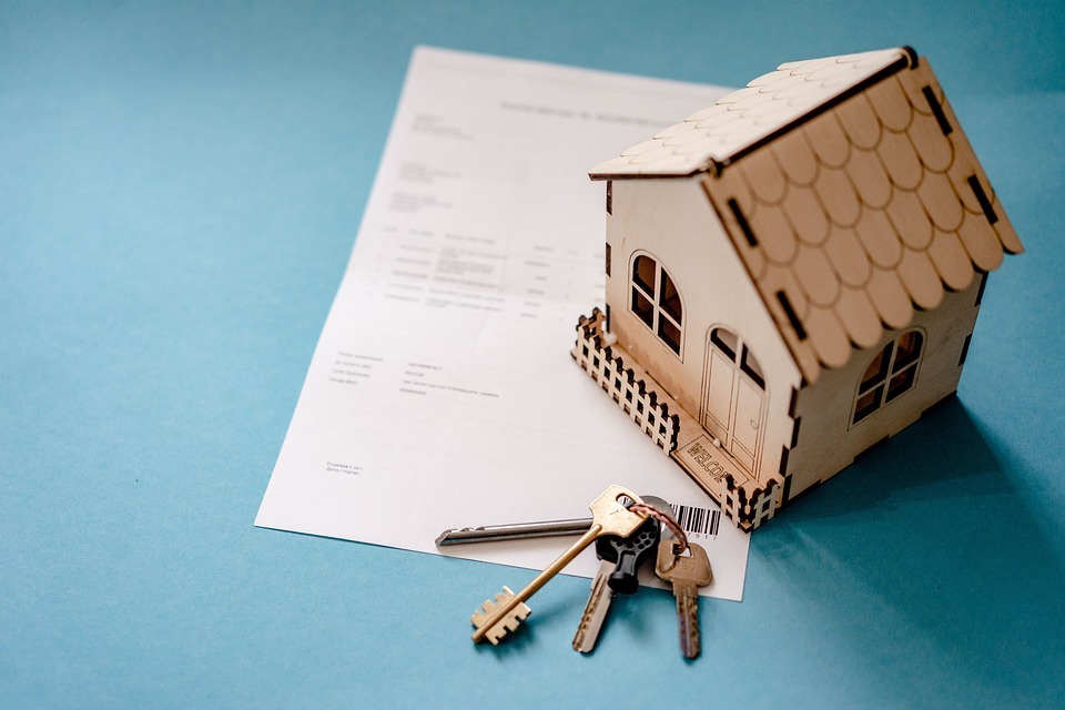 A small wooden model house sits on top of a document. A set of keys with a black keychain lies in the foreground on the document, which is placed on a blue background. It's an ideal visual for discussing Realtor Vs Real Estate Agent distinctions.